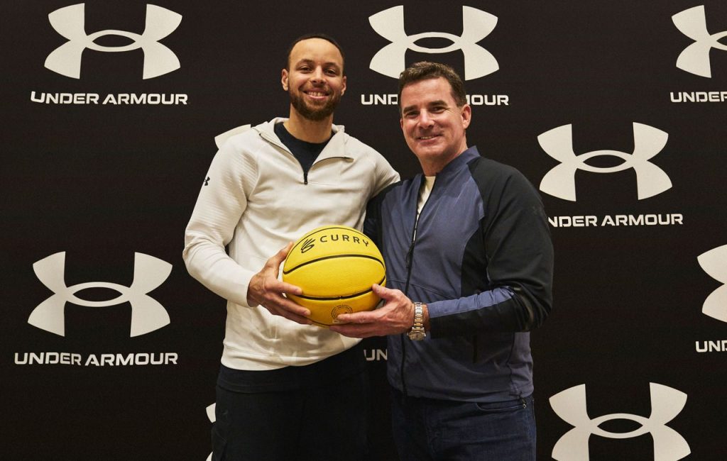 two men shown holding a yellow basketball in front of a wall with the Under Armour logo on it